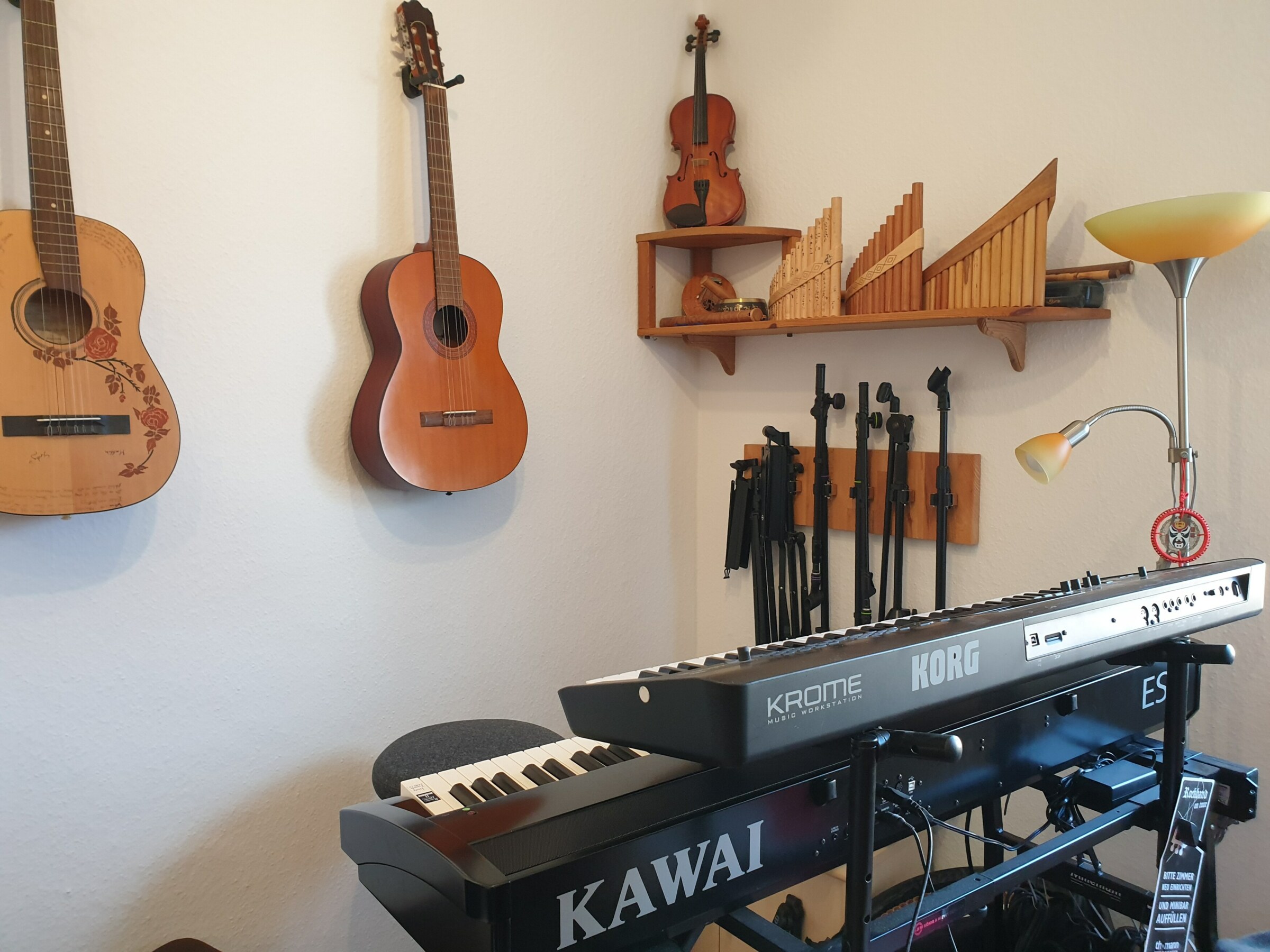 Foto einer Musikecke: Zwei digitale Klaviere auf einem Gestell übereinander. Im Hintergrund an der Wand hängen zwei Gitarren. An der anderen Wand hängen einige Stative. Auf einem Regal stehen Panflöten und eine Geige
