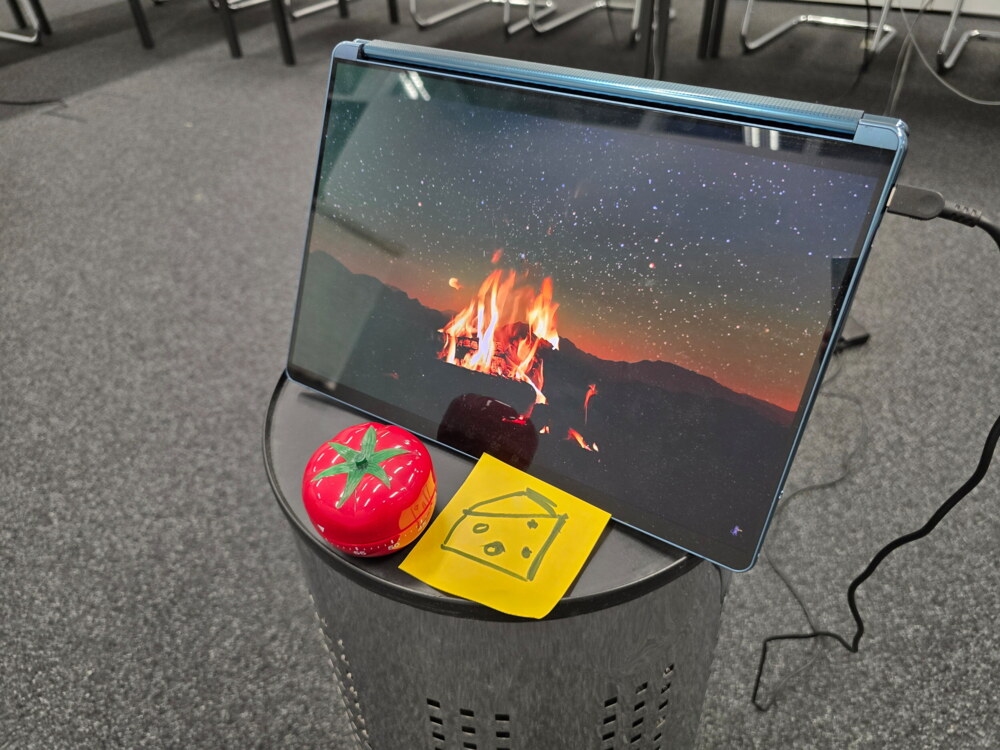 Laptop-Bildschirm zeigt ein Lagerfeuer unter Sternenhimmel, daneben eine tomatenförmige Kurzzeituhr und ein gelber Notizzettel mit gezeichnetem Käsestück, aufgestellt auf einem Hocker inmitten eines leeren Stuhlkreises.
