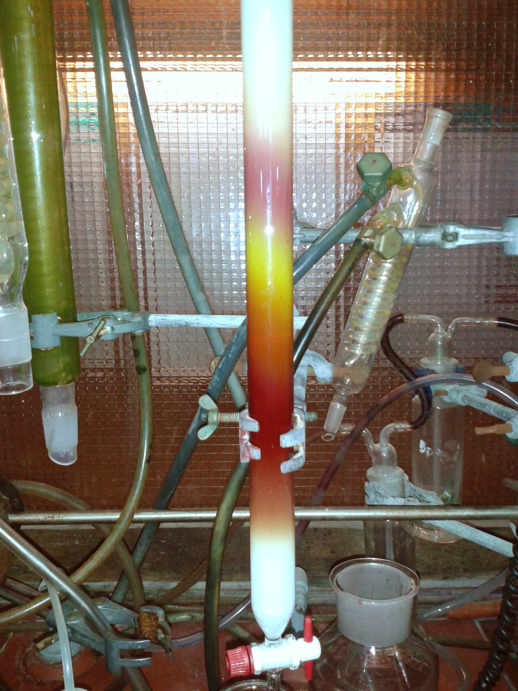 Chemie-Apparaturen. An einem Metallgestell ist ein Glaszylinder gefüllt mit weißem Sand befestigt. In dieser Säule sieht man einen Farbverlauf von braun über gelb zu lila.