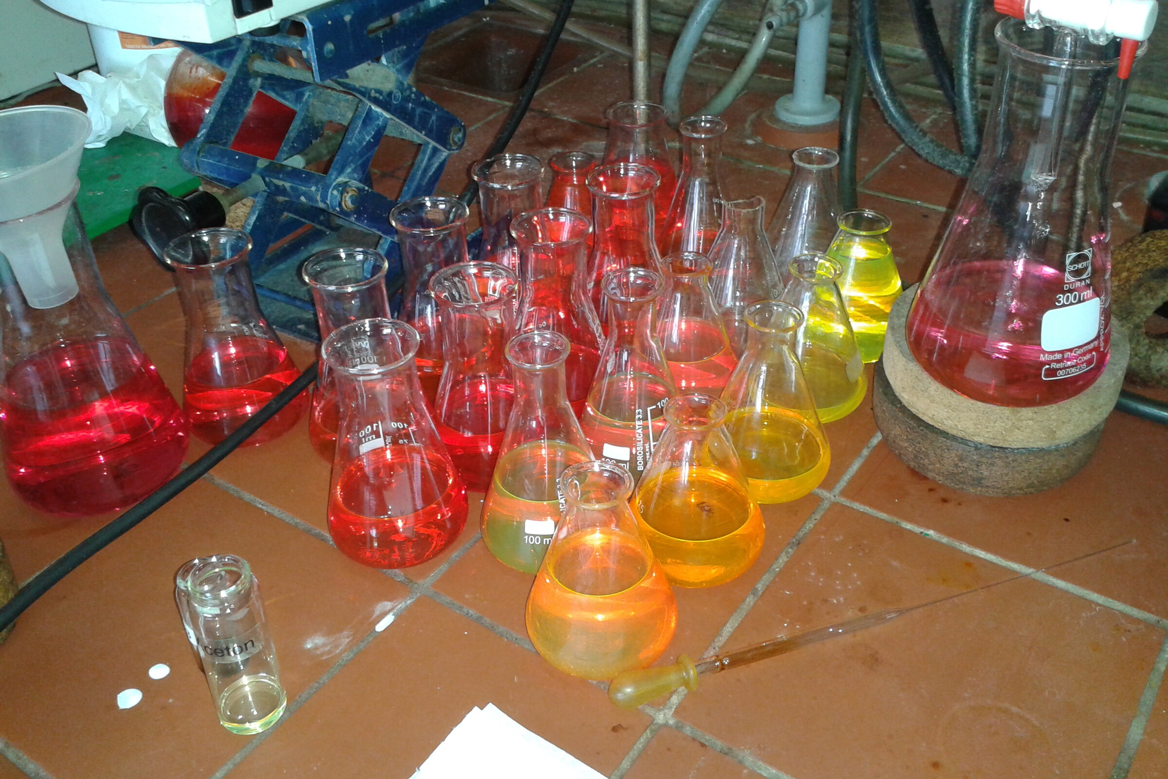 Auf einem Terrakotta-Fliesenboden im chemischen Labor stehen Glaskolben mit Flüssigkeit. Jeder Kolben hat eine andere Farbe; der Farbverlauf geht von Gelb über Orange bis hin zu Rot. Im Hintergrund stehen chemische Gerätschaften