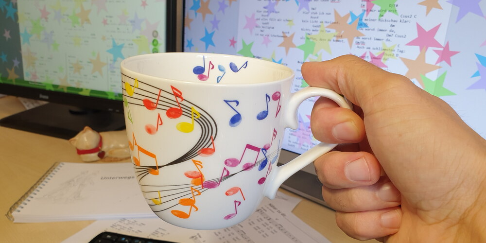 Ich halte eine Tasse mit bunten aufgedruckten Noten in der Hand. Im Hintergrund ist ein Schreibtisch mit Zetteln, einer Tastatur und Monitoren. Auf den Monitoren sind bunte Sternchen abgebildet