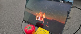 Laptop-Bildschirm zeigt ein Lagerfeuer unter Sternenhimmel, daneben eine tomatenförmige Kurzzeituhr und ein gelber Notizzettel mit gezeichnetem Käsestück, aufgestellt auf einem Hocker inmitten eines leeren Stuhlkreises.