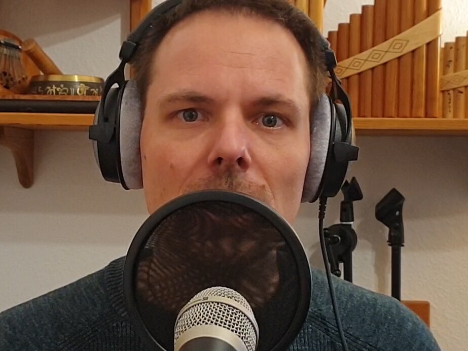 Ich singe in ein Mikrofon. Ein Netz als Ploppschutz ist zwischen Mikrofon und Mund gespannt. Ich habe Kopfhörer auf. Im Hintergrund stehen Panflöten und eine Klangschale auf dem Regal.
