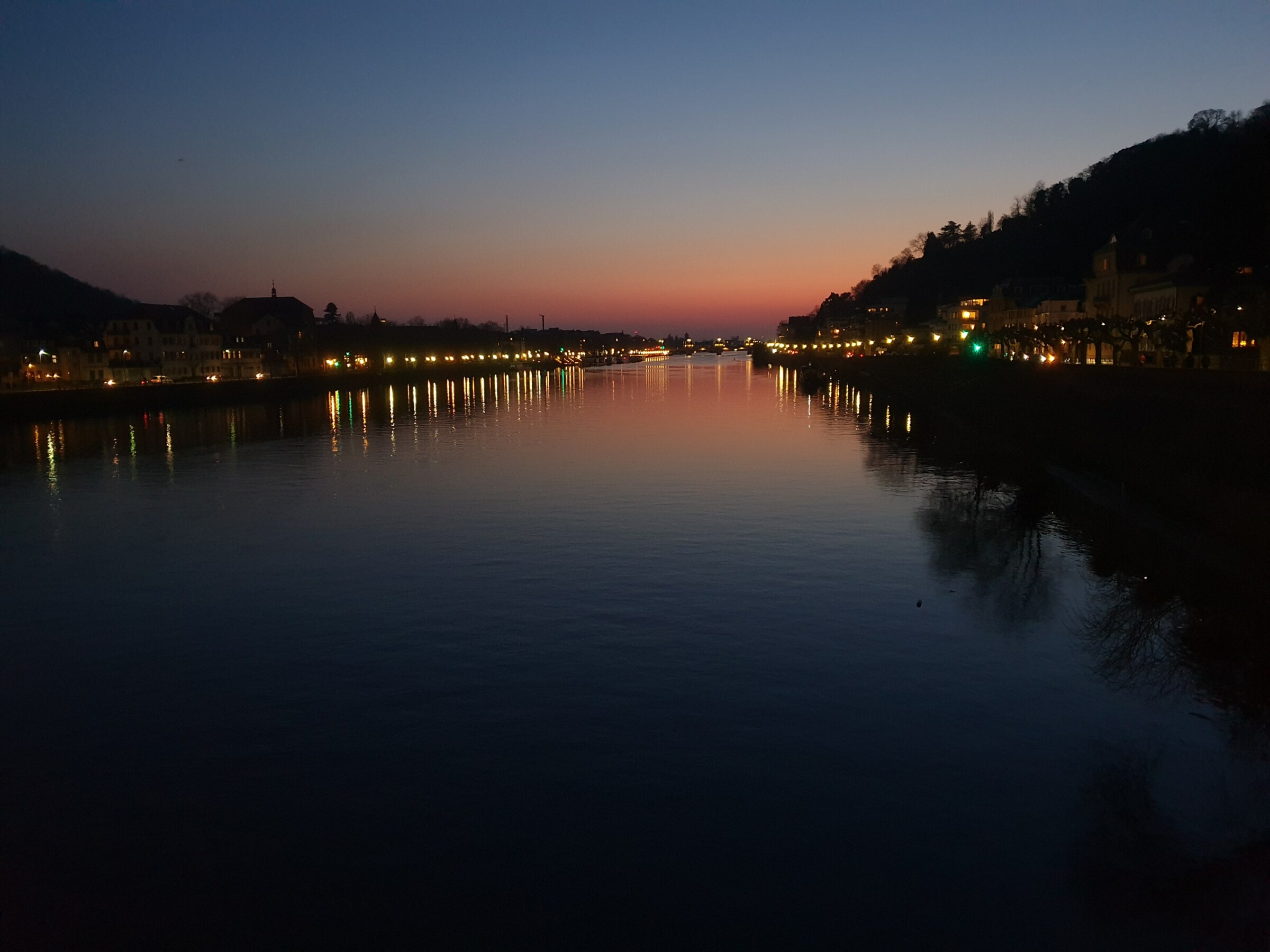 Ein Fluss bei Nacht. Links und rechts am Flußufer leuchten Lichter. Am Horizont strahlt das Abendrot der untergegangenen Sonne. Alles spiegelt sich auf der glatten Wasseroberfläche