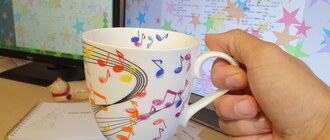 Ich halte eine Tasse mit bunten aufgedruckten Noten in der Hand. Im Hintergrund ist ein Schreibtisch mit Zetteln, einer Tastatur und Monitoren. Auf den Monitoren sind bunte Sternchen abgebildet