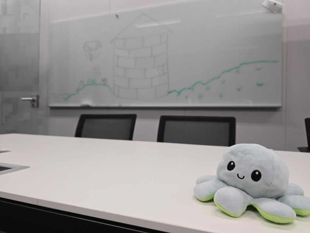 Konferenztisch mit einem Plüsch-Oktopus im Vordergrund, im Hintergrund ein Whiteboard mit Skizze: ein gemauerter Turm und zwei ansteigende Kurvenlinien mit kleinen Vögeln, umgeben von Stühlen im leeren Meetingraum.