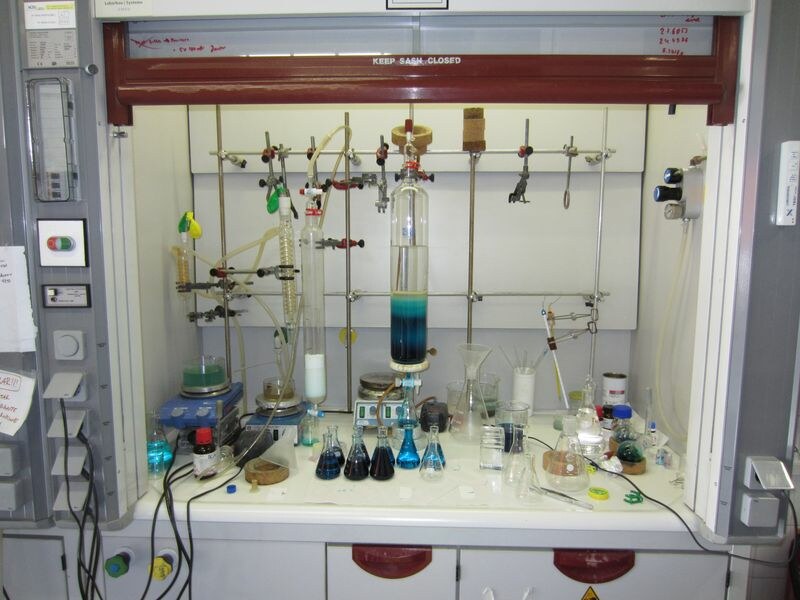 In einem geöffneten chemischen Abzug mit vielen Gerätschaften steht eine Säulenchromatographie. Unten kommt blaue Flüssigkeit raus und wird in Kolben aufgefangen.