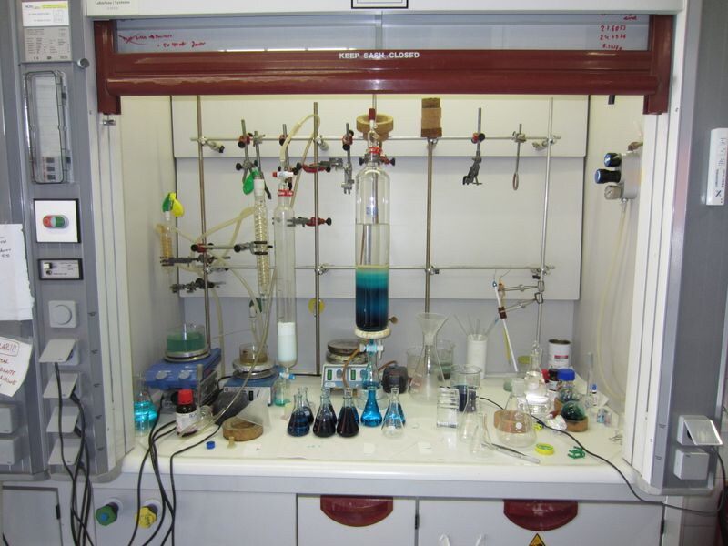 In einem geöffneten chemischen Abzug mit vielen Gerätschaften steht eine Säulenchromatographie. Unten kommt blaue Flüssigkeit raus und wird in Kolben aufgefangen.