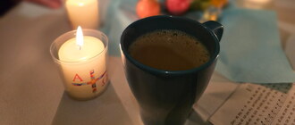 Eine Tasse Kaffee steht auf dem Tisch, neben einer brennenden Osterkerze. Im Hintergrund sieht man verschwommen eine Schale mit bunten Ostereiern, einem Liedzettel und weiteren Kerzen