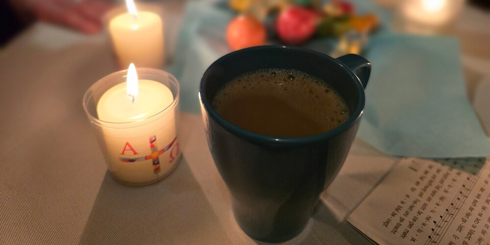 Eine Tasse Kaffee steht auf dem Tisch, neben einer brennenden Osterkerze. Im Hintergrund sieht man verschwommen eine Schale mit bunten Ostereiern, einem Liedzettel und weiteren Kerzen