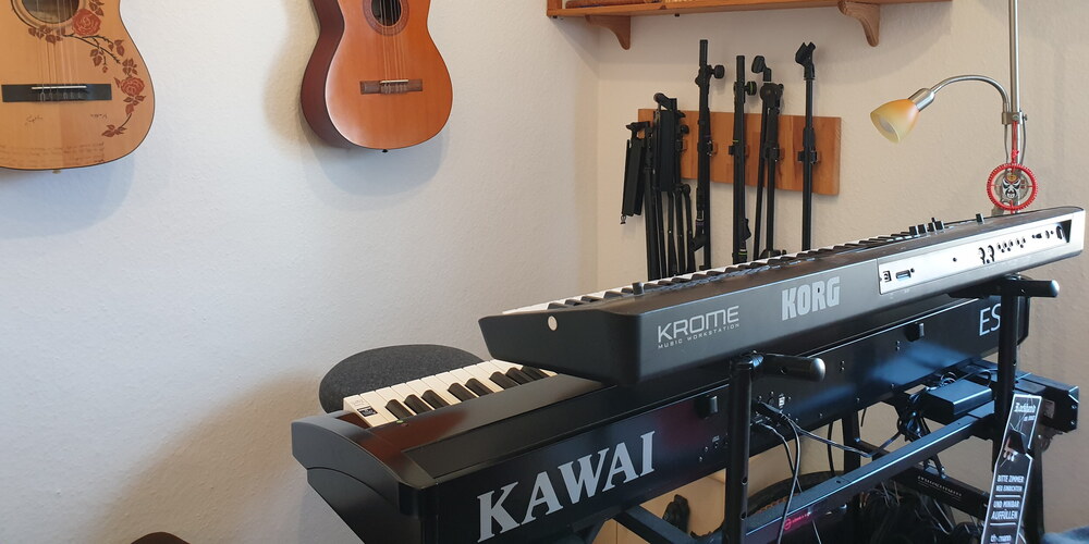 Foto einer Musikecke: Zwei digitale Klaviere auf einem Gestell übereinander. Im Hintergrund an der Wand hängen zwei Gitarren. An der anderen Wand hängen einige Stative. Auf einem Regal stehen Panflöten und eine Geige