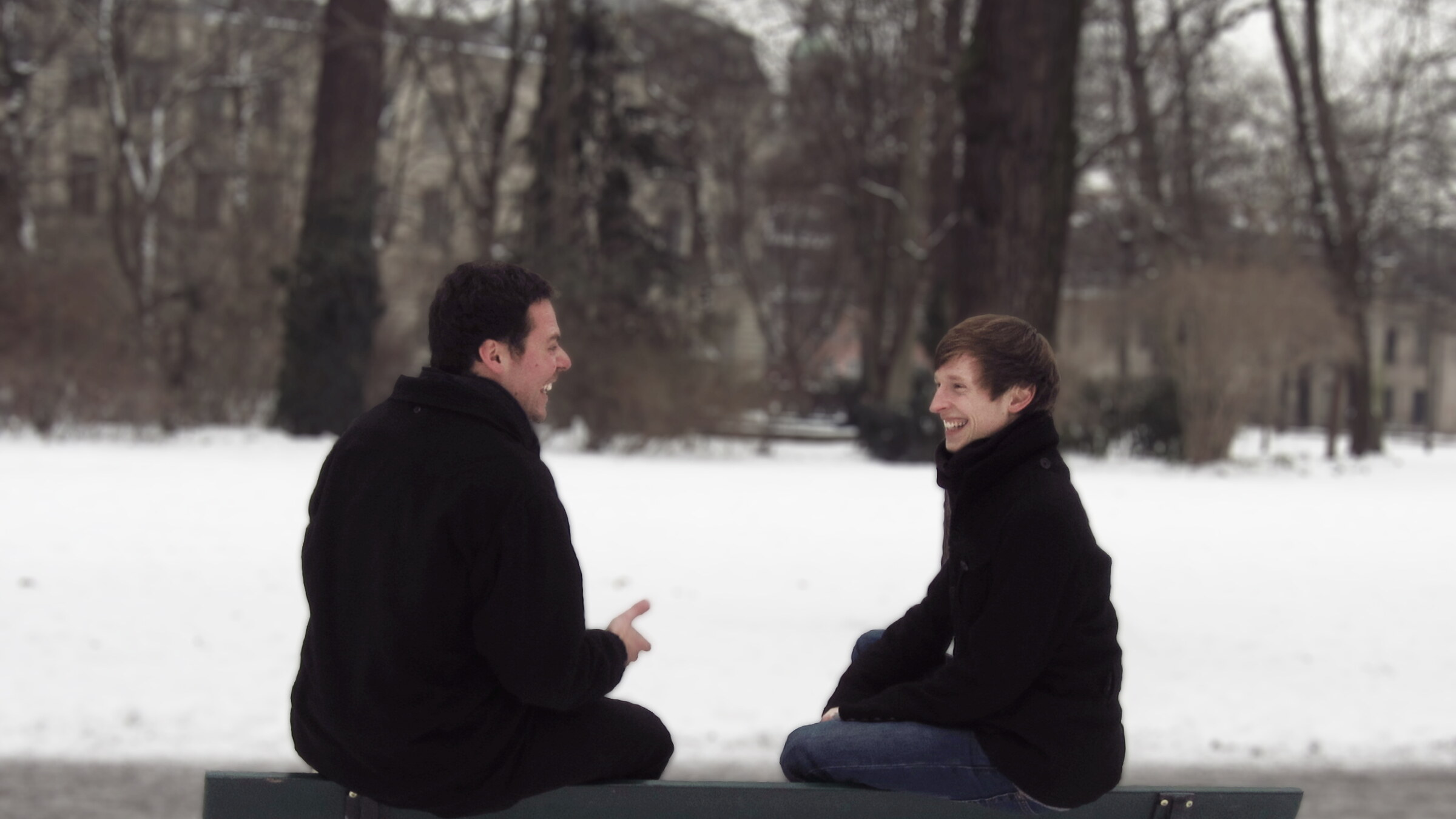 Zwei junge Männer sitzen auf einer Bank und unterhalten sich vor einer verschneiten Winderlandschaft.